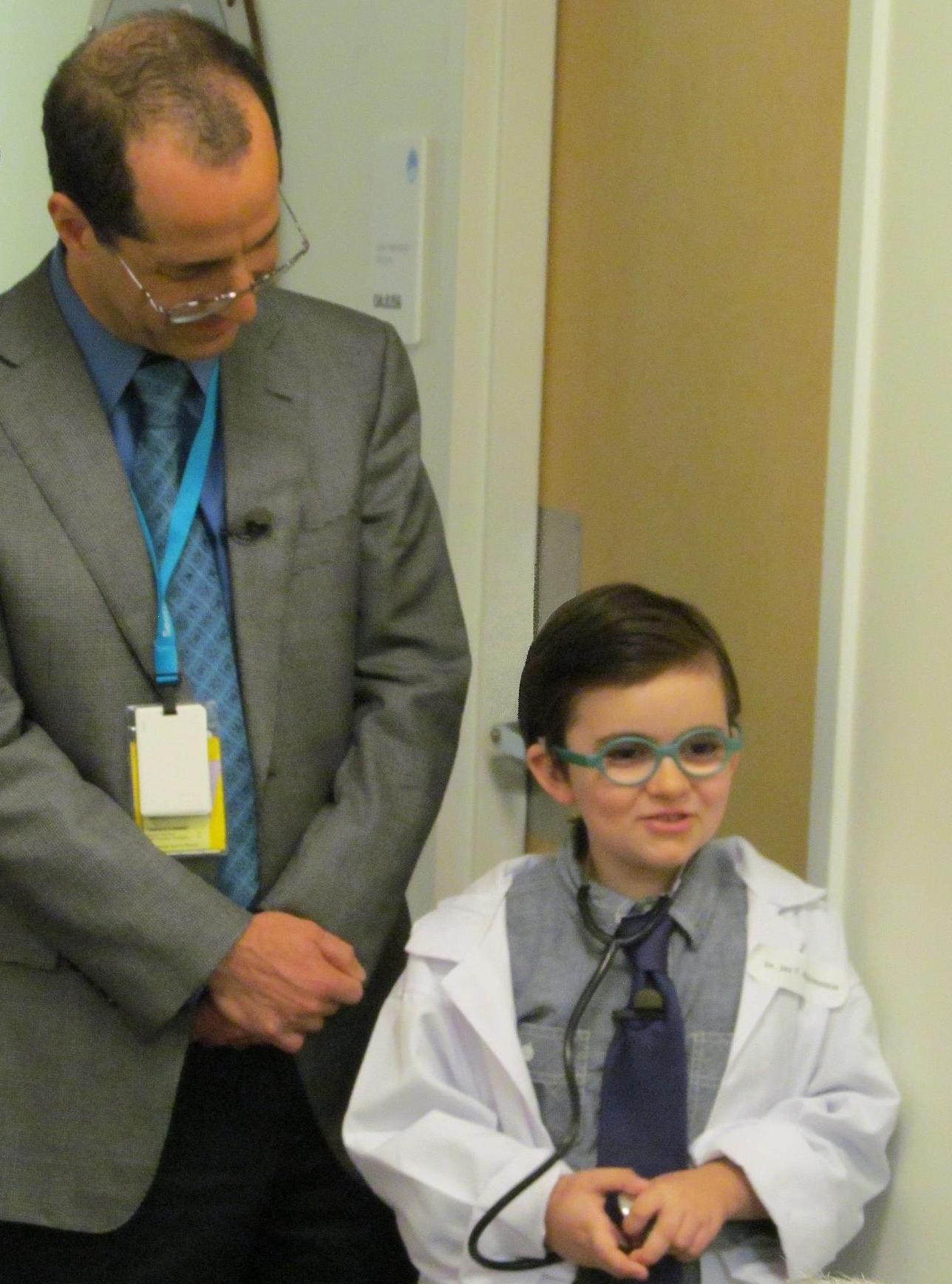 Photo of a tall man in glasses wearing a tie looking down at a young boy wearing play glasses, a tie, a white coat, and a stethoscope.