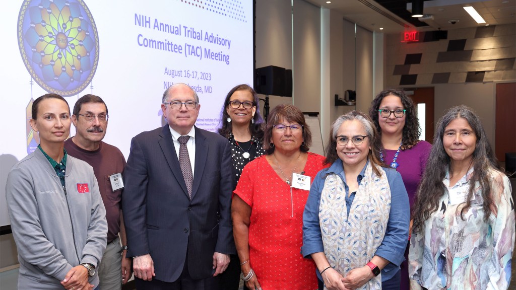 Group of people standing in front of a screen with "NIH Annual Tribal Committee (TAC) Meeting" displayed