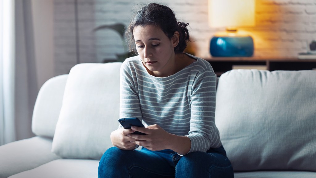 A distraught woman sits on a couch interacting with her phone