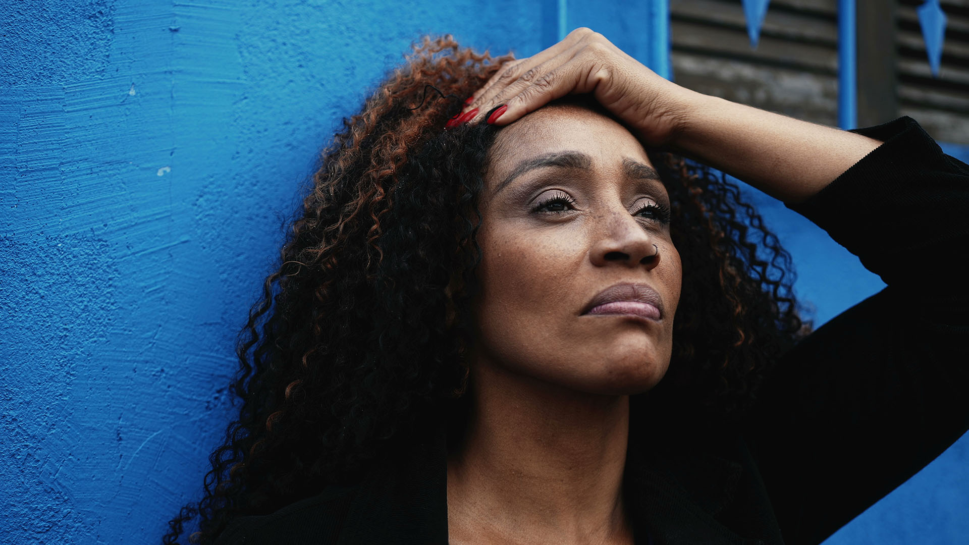 A black woman leans against a wall with her hand on her head looking anxious