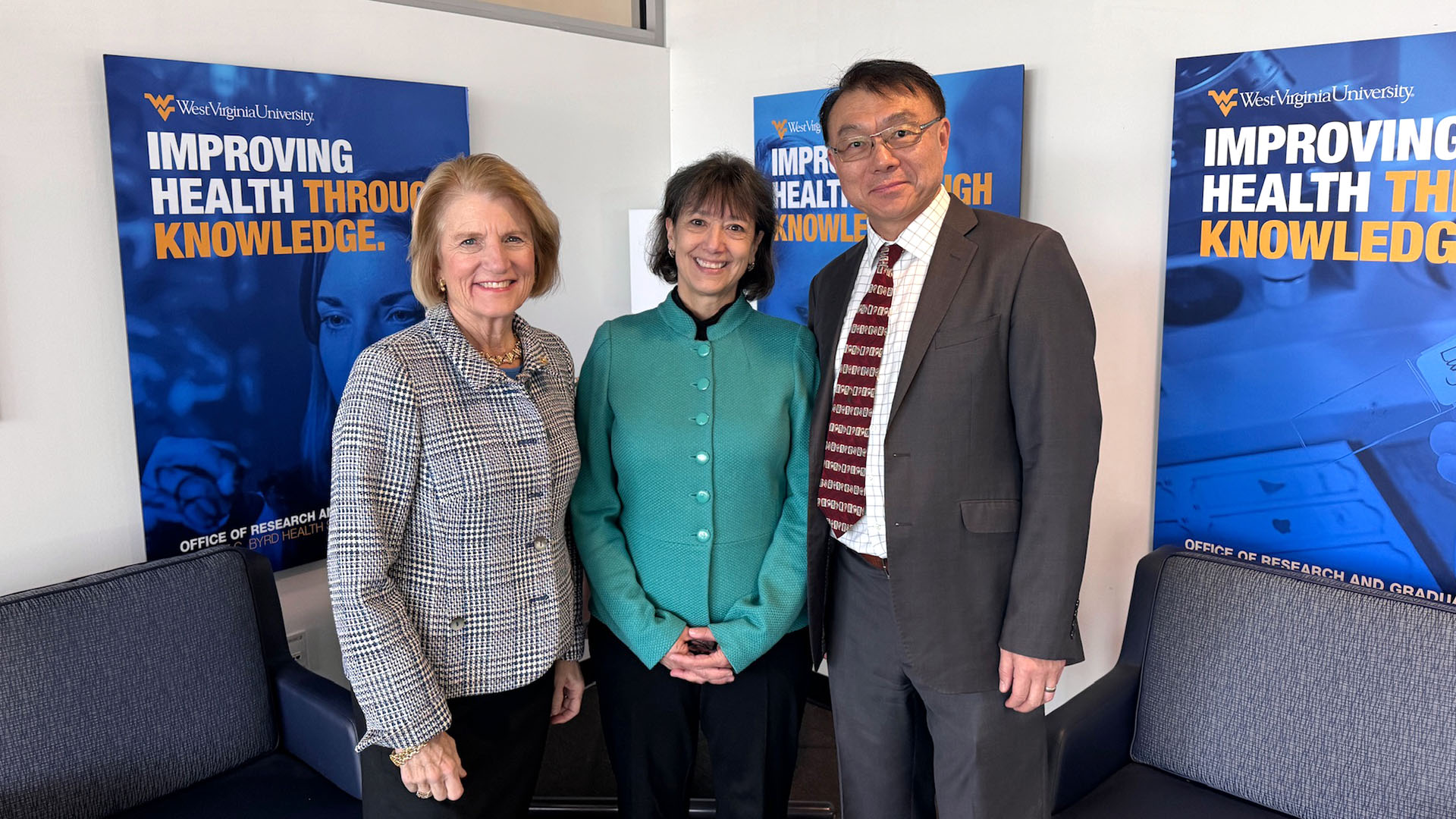 NIH Director Monica Bertagnolli with Senator Shelley Moore Capito, and Dr. Ming Lei, Vice Dean of Research at the WVU School of Medicine standing in front of posters that read 