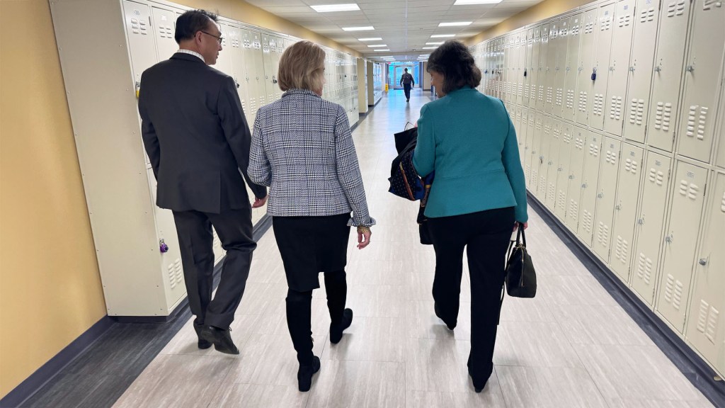 A man and two women walking away down a hall lined with lockers